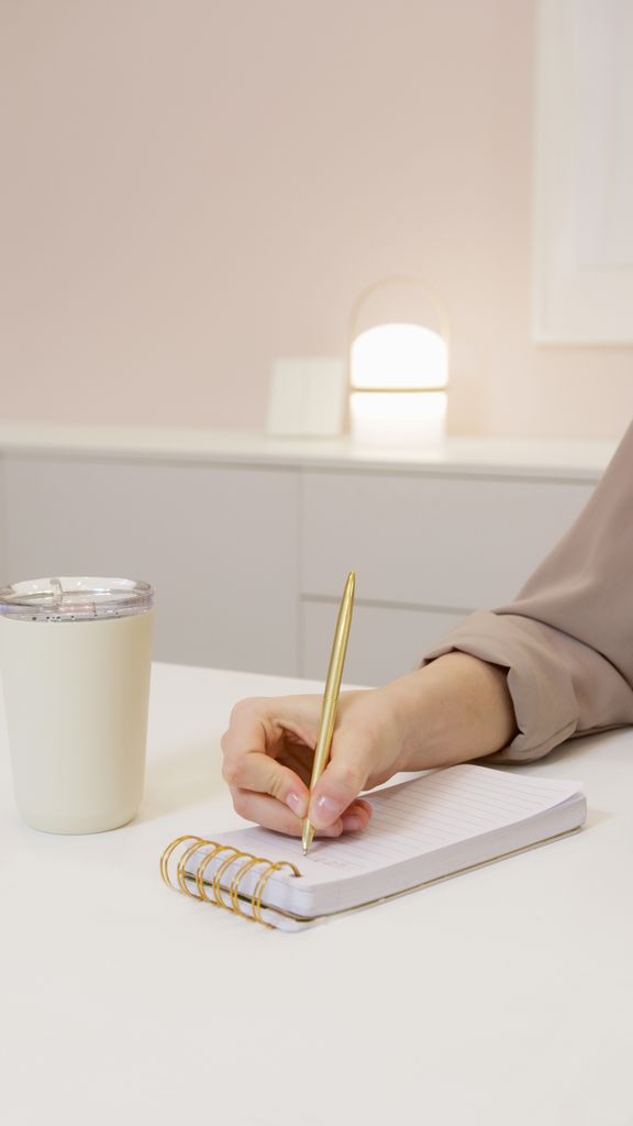 A close-up of a hand holding a pen, writing notes in a notebook on a minimalist office desk.